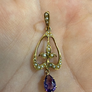 May include: A gold-toned pendant featuring a teardrop-shaped amethyst gemstone suspended below a decorative openwork design. The pendant is adorned with small pearls and has a delicate, ornate appearance, suitable for jewelry.