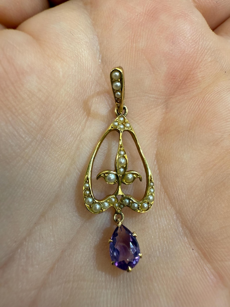 May include: A gold-toned pendant featuring a teardrop-shaped amethyst gemstone suspended below a decorative openwork design. The pendant is adorned with small pearls and has a delicate, ornate appearance, suitable for jewelry.
