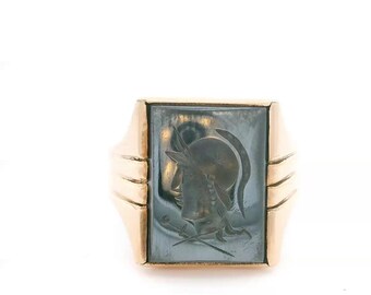 May include: A gold ring featuring a rectangular, dark gray hematite intaglio set in the center. The stone is carved with the profile of a helmeted figure and a sword. The band has a polished gold finish with three horizontal grooves on each side.