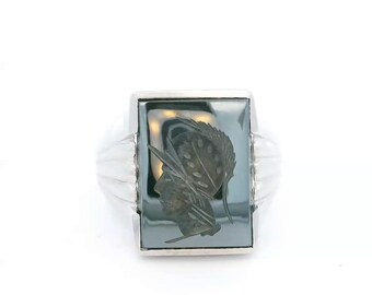 May include: A silver ring featuring a rectangular, dark gray hematite stone set in the center. The stone is engraved with a profile view of a warrior wearing a plumed helmet. The ring band has a ribbed design on both sides of the setting.