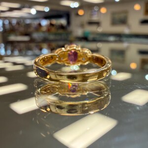 May include: A gold ring with a central oval pink gemstone and smaller clear stones. The ring has a detailed, ornate design and is set against a reflective surface, showcasing its intricate craftsmanship and elegant style.