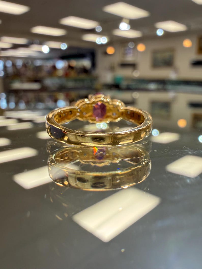 May include: A gold ring with a central oval pink gemstone and smaller clear stones. The ring has a detailed, ornate design and is set against a reflective surface, showcasing its intricate craftsmanship and elegant style.