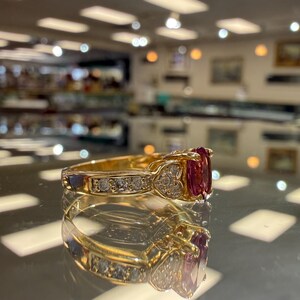 May include: A gold ring with a large, oval-cut, red gemstone flanked by smaller diamonds. The band features additional diamonds and heart-shaped accents. The ring is displayed on a reflective surface.