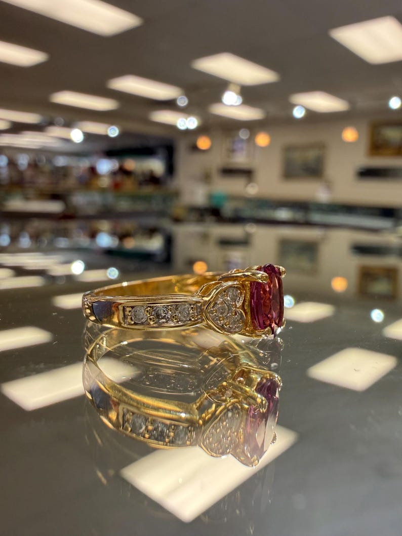 May include: A gold ring with a large, oval-cut, red gemstone flanked by smaller diamonds. The band features additional diamonds and heart-shaped accents. The ring is displayed on a reflective surface.