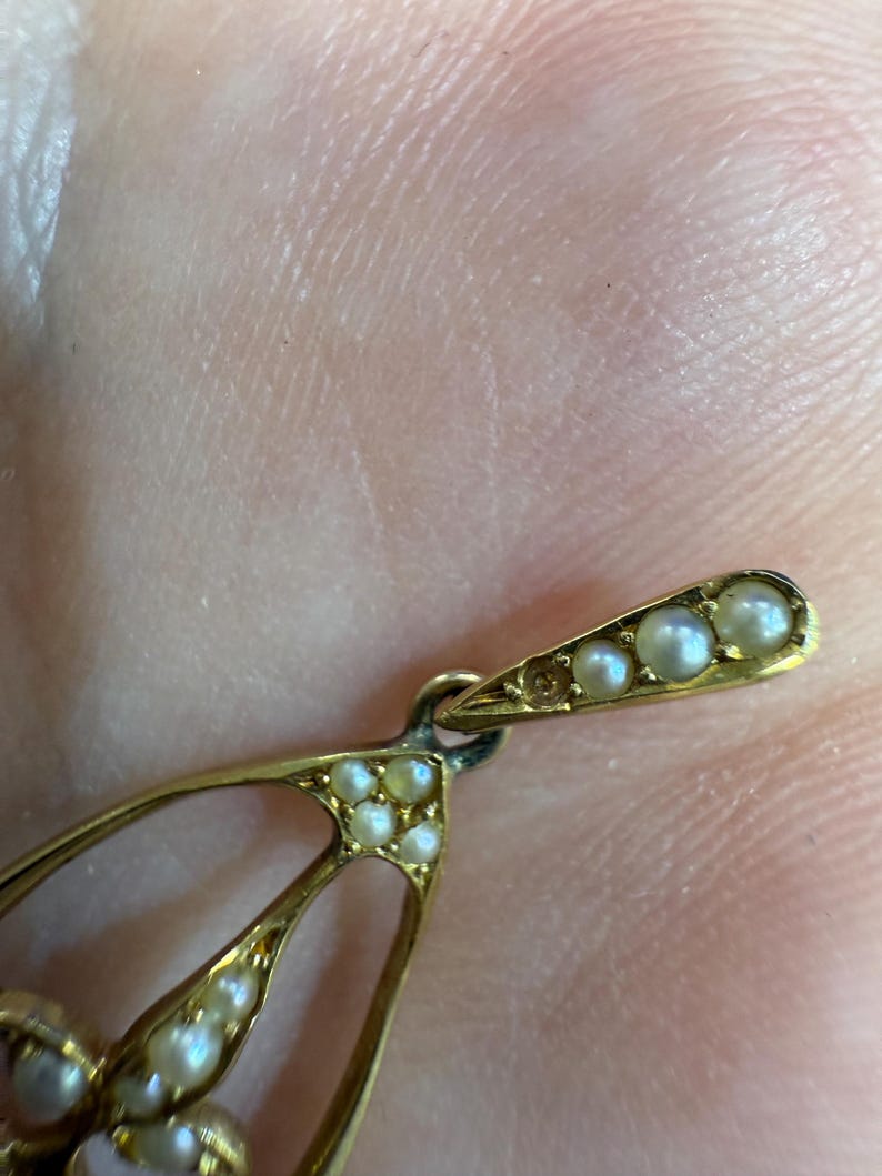 May include: Close-up of a gold-toned pendant with a teardrop design. The pendant is adorned with small, round pearls. The top section features a row of pearls, and the main body has multiple pearls.