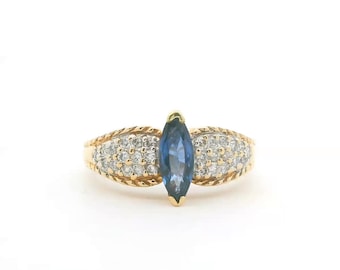 May include: A gold ring featuring a central marquise-cut blue gemstone flanked by two clusters of small, clear round stones. The band has a textured, rope-like edge detail on both sides of the central setting, creating a distinct visual contrast.