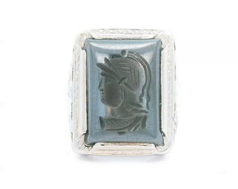 May include: A silver ring featuring a rectangular, grey, polished stone inlay. The stone is carved with a profile relief of a figure wearing a helmet with a crest. The metal setting has a textured, decorative border surrounding the central stone.
