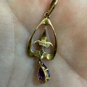 May include: A gold pendant with a teardrop-shaped amethyst gemstone. The pendant has an openwork design with a floral motif and a loop for a chain. The metal is a warm gold color, and the gemstone is a deep purple.