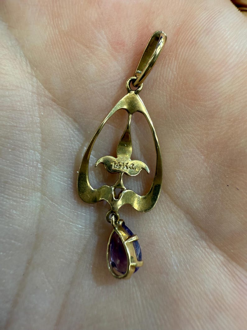 May include: A gold pendant with a teardrop-shaped amethyst gemstone. The pendant has an openwork design with a floral motif and a loop for a chain. The metal is a warm gold color, and the gemstone is a deep purple.