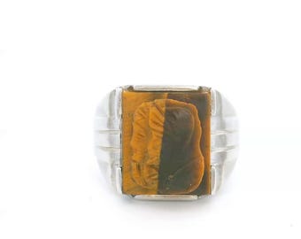 May include: A silver ring featuring a rectangular tiger's eye gemstone set in the center. The stone is carved with a profile portrait of a person, displaying natural brown and golden bands. The metal band has a smooth, polished finish.