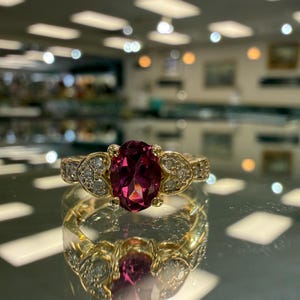 May include: A gold ring with a large oval, deep pink gemstone as the centerpiece. The ring is accented with smaller, clear gemstones and intricate detailing on the band. The ring is displayed on a reflective surface.