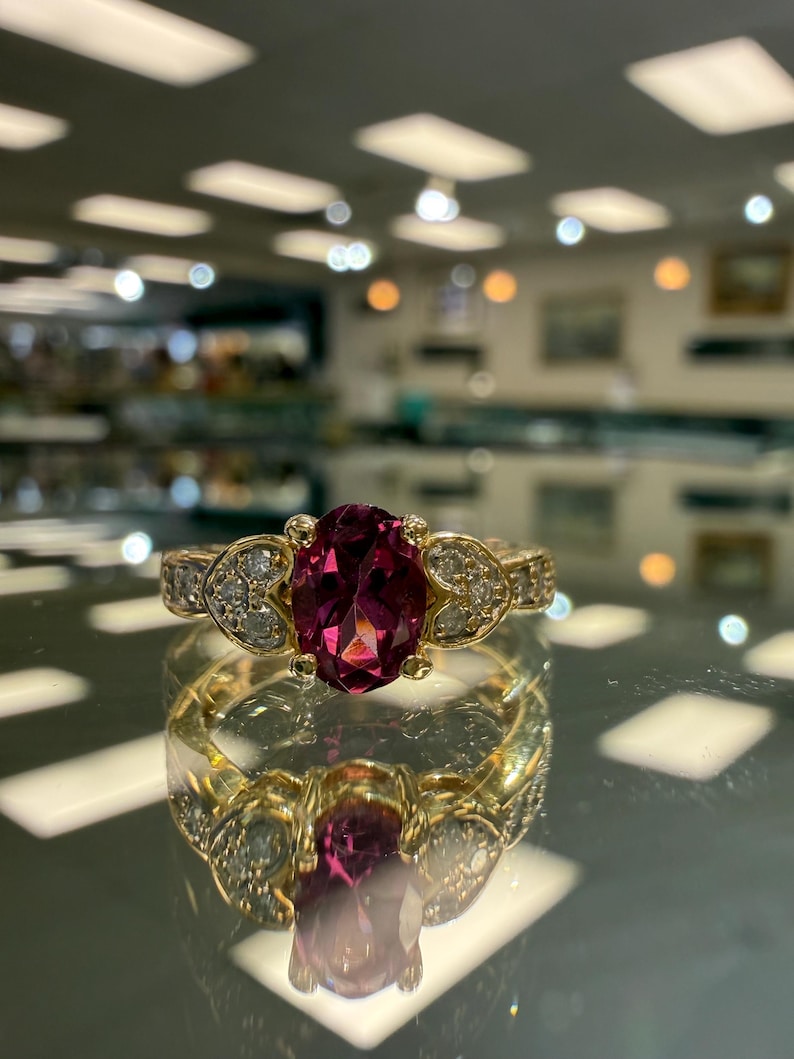 May include: A gold ring with a large oval, deep pink gemstone as the centerpiece. The ring is accented with smaller, clear gemstones and intricate detailing on the band. The ring is displayed on a reflective surface.