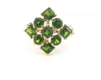 May include: A gold ring featuring a cluster of nine green gemstones arranged in a diamond-shaped pattern. The design includes four square-cut stones and five round-cut stones, all set in a yellow gold metal band with small prongs.