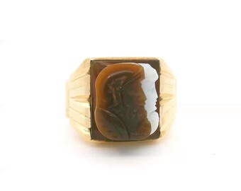 May include: A gold ring featuring a rectangular cameo centerpiece. The cameo displays a dual-profile carving with a brown, helmeted figure on the left and a white, smooth-featured figure on the right, set within a polished gold band.