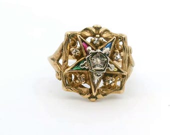 May include: A gold ring featuring an ornate square frame with filigree details. At the center is a five-pointed star with colored enamel in red, blue, and green, accented by small clear stones and a central diamond-like setting in white metal.
