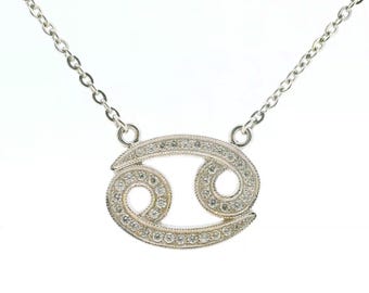 May include: A silver-toned necklace featuring a Cancer zodiac symbol pendant. The pendant is encrusted with numerous small, clear, sparkling stones along its entire surface and is attached to a delicate, thin silver-toned metal chain.