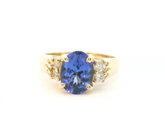 May include: A gold ring featuring a large, oval-cut blue gemstone set in the center, flanked by two small, round, clear stones on each side. The band is polished yellow gold, and the setting uses prongs to secure the central blue stone.