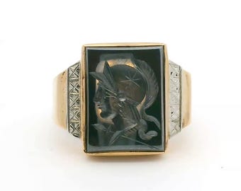 May include: A gold ring featuring a rectangular black stone inlay with a carved profile of a figure wearing a plumed helmet. The band has silver-toned side panels with a cross-hatched pattern, contrasting with the polished gold shank.