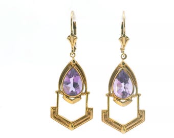 May include: A pair of gold drop earrings featuring a pear-shaped purple amethyst gemstone set within a gold frame, suspended from a lever-back closure. The design includes a geometric gold structure hanging below each faceted purple stone.