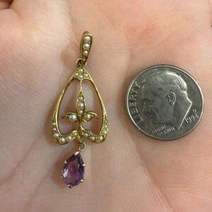 May include: A gold-toned pendant with a teardrop-shaped amethyst gemstone and small pearls. The pendant has an openwork design with a floral motif and is shown next to a U.S. dime for size comparison. Jewelry.
