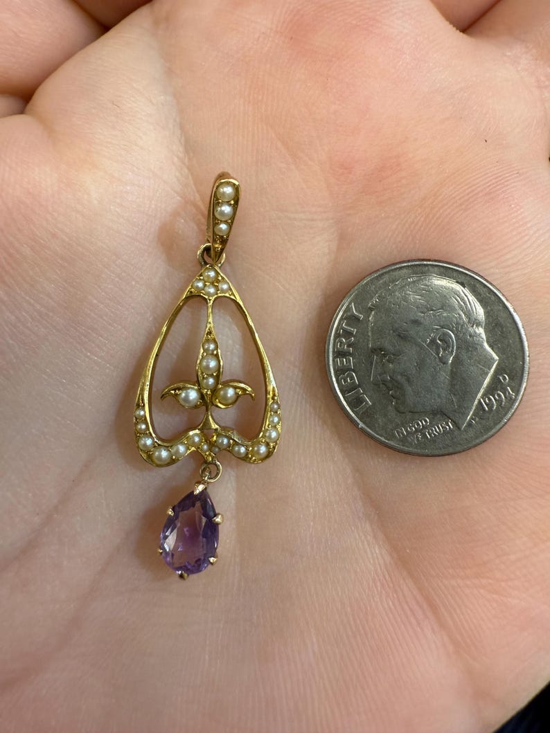 May include: A gold-toned pendant with a teardrop-shaped amethyst gemstone and small pearls. The pendant has an openwork design with a floral motif and is shown next to a U.S. dime for size comparison. Jewelry.