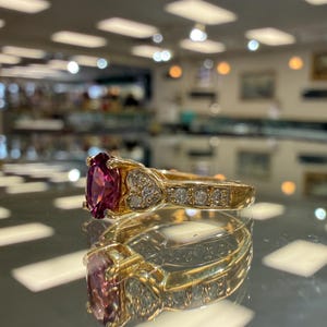 May include: A gold ring with a large, oval-cut, deep pink gemstone as the centerpiece. The band features small, clear accent stones and heart-shaped designs. The ring is displayed on a reflective surface.