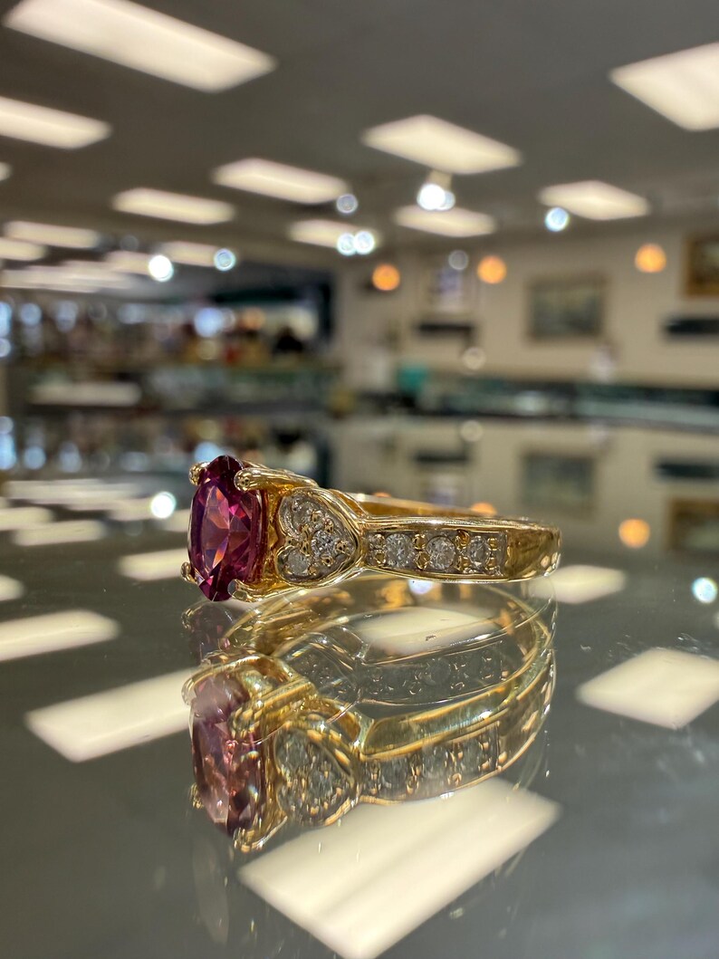 May include: A gold ring with a large, oval-cut, deep pink gemstone as the centerpiece. The band features small, clear accent stones and heart-shaped designs. The ring is displayed on a reflective surface.