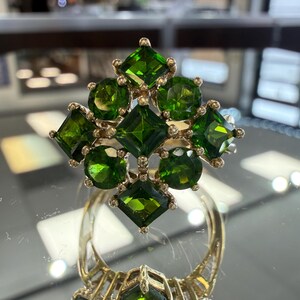 May include: A gold ring featuring a cluster of green gemstones. The ring's design includes square and round-cut stones, arranged in a floral or starburst pattern. The ring is displayed on a reflective surface.