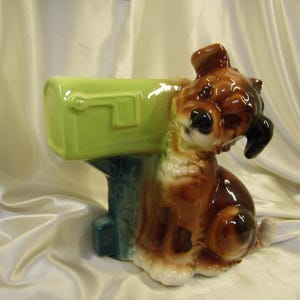 May include: A ceramic dog figurine with a mailbox planter. The dog is brown and white with black accents, sitting next to a light green mailbox. The mailbox is supported by a blue base. The figurine has a glossy finish.