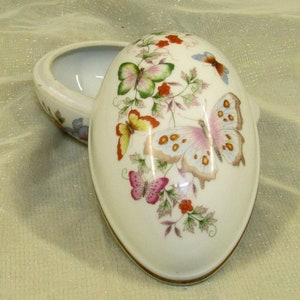 May include: An egg-shaped trinket box with a hinged lid. The off-white porcelain box is decorated with colorful butterflies and floral accents. The box has a gold-colored trim around the base and lid.