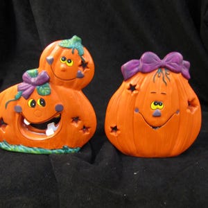 May include: Two orange ceramic pumpkin figurines with painted faces, purple bows, and star-shaped cutouts. The left figurine features a stacked design with two pumpkins, while the right figurine is a single pumpkin with a wide, smiling expression.
