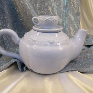 May include: A light blue ceramic teapot with a matching teacup and saucer on top. The teapot has a curved handle and spout, and the surface is decorated with embossed floral patterns. The teacup is small and delicate, with a scalloped rim.