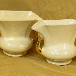 May include: Two cream-colored vases with gold handles. The vases have a flared, geometric shape and a pedestal base. The gold handles curve gracefully. The vases are displayed on a gold-colored surface. These decorative vases are suitable for home decor.