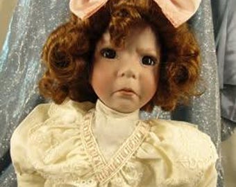 May include: A porcelain doll with brown curly hair, wearing a white dress with lace trim and a pink bow.