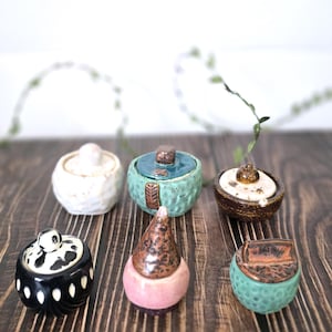 May include: A collection of small, handcrafted ceramic jars with lids. The jars are in various colors, including white, turquoise, black, and pink, and feature unique textures and patterns. The lids have different shapes and designs, adding to the artistic appeal.