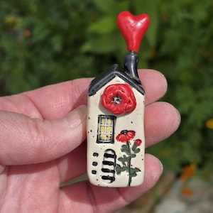 May include: A small ceramic house figurine featuring a cream-colored base, a black roof, and a chimney topped with a red heart. The front displays a raised red poppy flower, a painted yellow window, a black door, and a painted red poppy plant.