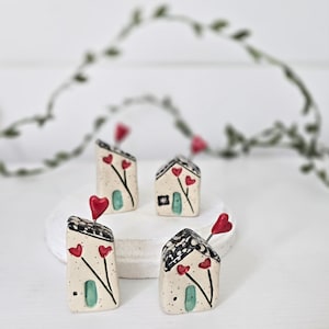 May include: Four small ceramic houses with black and white patterned roofs, turquoise doors, and red heart accents. Each house has a red heart on a wire extending from the roof. The houses are on a white surface.