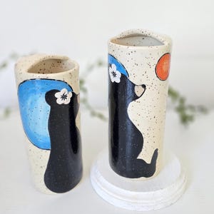 May include: Two speckled ceramic vases with a bear design. One vase features a blue circle, the other an orange circle. Each vase has a black bear silhouette with a white flower. The vases are on a white surface.