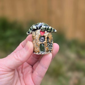 May include: A miniature ceramic house with a textured, tan-colored body, a green and black roof, and a red heart detail. The house features a small green door and decorative elements.