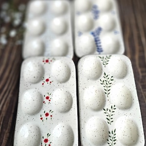May include: Four white ceramic egg trays with speckled surfaces. Two trays have floral designs in red and green, while the other two feature blue floral patterns. Each tray holds six eggs.