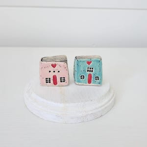 以下が含まれることがあります： 薄いピンクと薄いブルーの小さな陶器製の家の置物が2つ。それぞれ赤いドアの上に赤いハート、黒く塗られた窓、黒い線のディテールが施された屋根が特徴です。家は斑点のある仕上げで、滑らかな質感です。