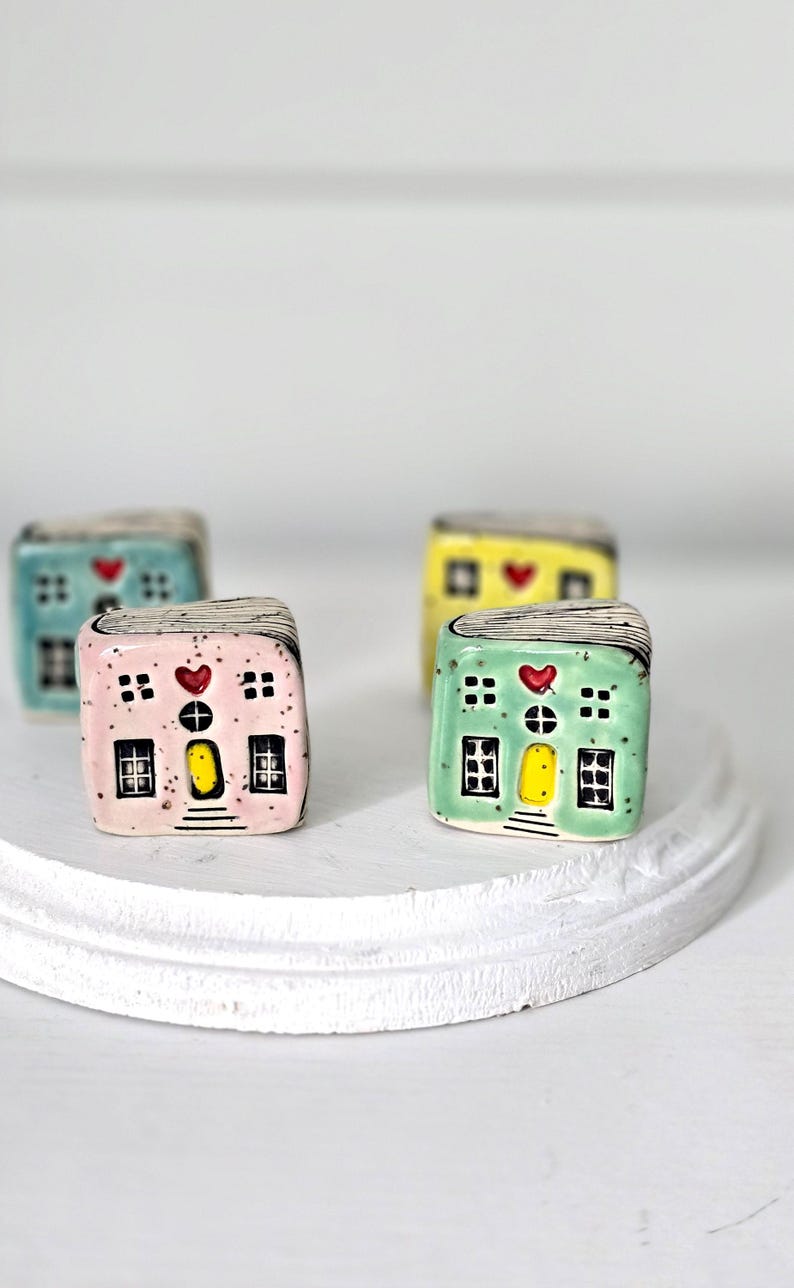 May include: Four small, cube-shaped ceramic house figurines in pastel pink, mint green, yellow, and blue. Each house features a yellow door, black-outlined windows, a red heart above the door, and a roof with black line details and speckles.