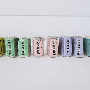 以下が含まれることがあります： 黄色、緑、ピンク、紫、青緑などのパステルカラーで彩られた、長方形の本の形をした10個の小さな陶器製オーナメント。各ピースの前面には、DREAMまたはINSPIREという黒い手押しスタンプの文字が刻まれています。