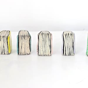 以下が含まれることがあります： 一列に並べられた6つの小さな陶器製の本の彫刻。各ピースは、黒い縦線が入った白いページ部分と、黒、黄色、ティール、ピンク、薄紫、薄緑の色の背表紙が特徴で、質感のある仕上がりになっています。