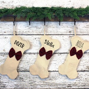 May include: Three bone-shaped Christmas stockings with names "DOZER", "STELLA", and "BELLA" in black script. Each stocking has a red and black plaid bow. They hang from a wooden shelf decorated with greenery.