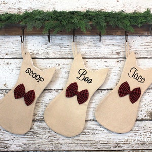 May include: Three beige cat-shaped Christmas stockings with red and black plaid bows. Each stocking has a name embroidered in black: "Scoop", "Boo", and "Taco". They hang from a wooden shelf with greenery.