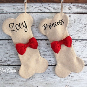 May include: Two bone-shaped Christmas stockings made of tan burlap, each with a red bow. One stocking is labeled "Zoey" and the other "Princess" in black script. The stockings hang from a wooden beam.