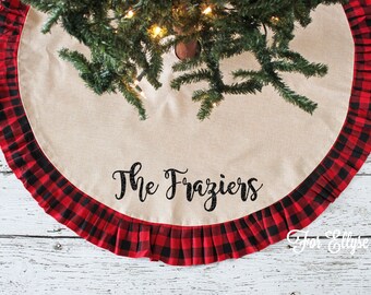 May include: A round Christmas tree skirt with a natural linen center and a red and black buffalo plaid ruffle border. The text "The Fraziers" is printed in black script across the center. The skirt is under a Christmas tree.