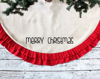 May include: A beige Christmas tree skirt with a red ruffled border. The words "MERRY CHRISTMAS" are printed in black across the center. The skirt is partially covered by a green Christmas tree.