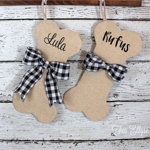 May include: Two beige dog bone-shaped Christmas stockings with black and white checkered bows. One stocking is labeled "Lula" and the other "Rufus". The stockings are hanging against a white wooden background.